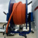 hydraulicplanet A close-up of a JC7 SEWER JETTING HOSE ASSEMBLY-100M COILS, orange and coiled on a blue metal reel mounted to a vehicle, with parked cars and buildings in the background.