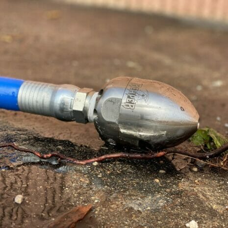 REAPER™ ROTATING JETTING NOZZLE 3/8" INLET, 24°-5 hydraulicplanet A close-up of a REAPER™ Rotating Jetting Nozzle 3/8" Inlet, 24°, attached to a blue hose and lying on wet concrete near a root.