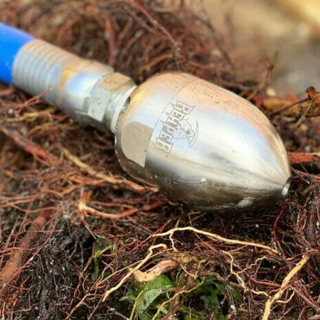 REAPER™ ROTATING JETTING NOZZLE 3/8" INLET, 24°-4 hydraulicplanet A REAPER™ ROTATING JETTING NOZZLE 3/8" INLET, 24° is connected to a blue hose and positioned among tangled roots and debris.